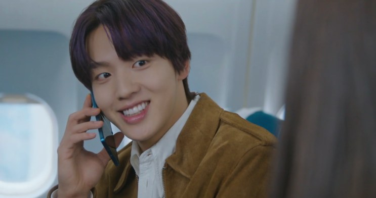 Jae-in falando ao telefone e sorrindo, dentro do avião