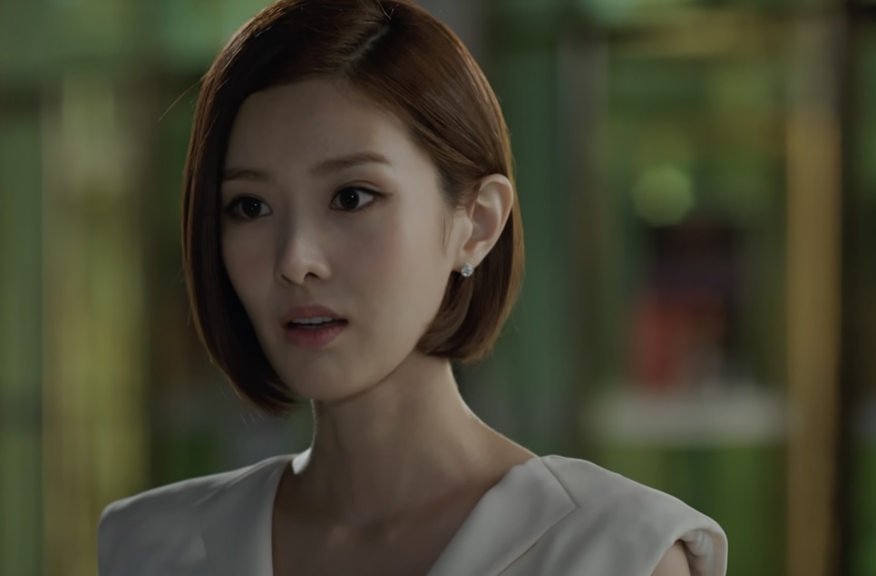 Tae Yi-ryung de blusa branca com expressão séria, em uma estufa de vidro