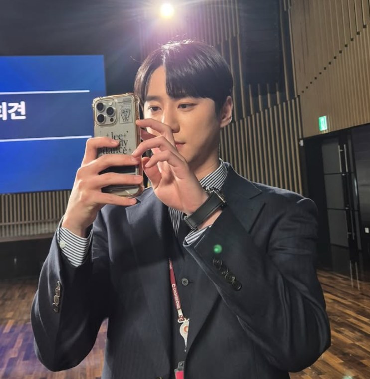 Lee Jun-young de terno, fotografando com o celular em frente a um painel de evento
