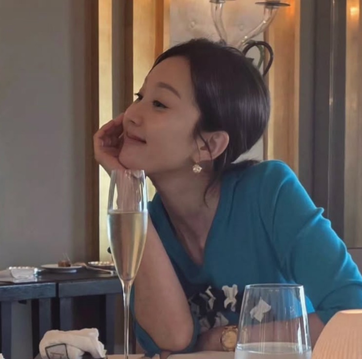 Yum Jung-ah hoje, de blusa azul, apoiando o rosto na mão à mesa